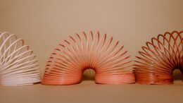 Trzy kolorowe sprężyny–zabawki ustawione obok siebie na jednolitym beżowym tle, rozłożone w charakterystycznym łukowatym kształcie.