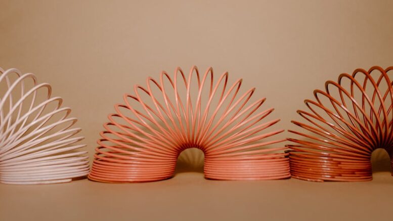 Trzy kolorowe sprężyny–zabawki ustawione obok siebie na jednolitym beżowym tle, rozłożone w charakterystycznym łukowatym kształcie.