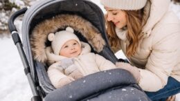 Matka na zimowym spacerze pochylająca się nad niemowlęciem leżącym w wózku, ubrane w ciepły kombinezon i czapkę, otoczone zimową scenerią