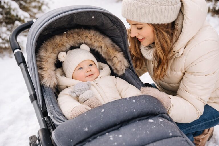 Matka na zimowym spacerze pochylająca się nad niemowlęciem leżącym w wózku, ubrane w ciepły kombinezon i czapkę, otoczone zimową scenerią